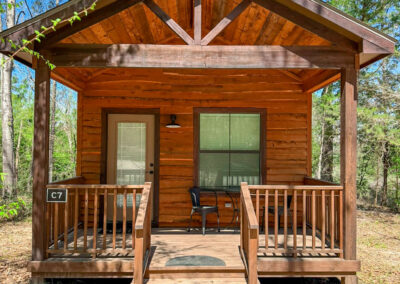Rustic wooden cabin with porch in forest setting, surrounded by trees, sunny sky. Ideal for nature retreat.