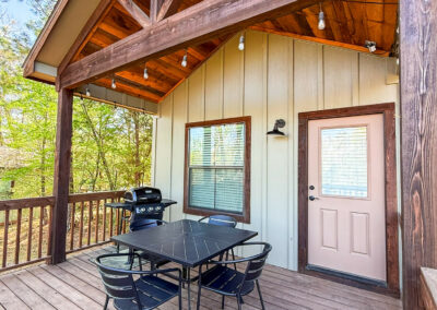 Rustic cabin porch with dining table and grill, surrounded by trees. Cozy outdoor living space.