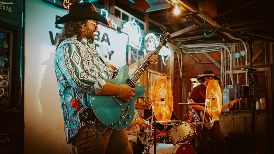 Live band performing in rustic venue with guitarist, neon lights, drums, and cowboy hats, creating an exciting musical atmosphere.
