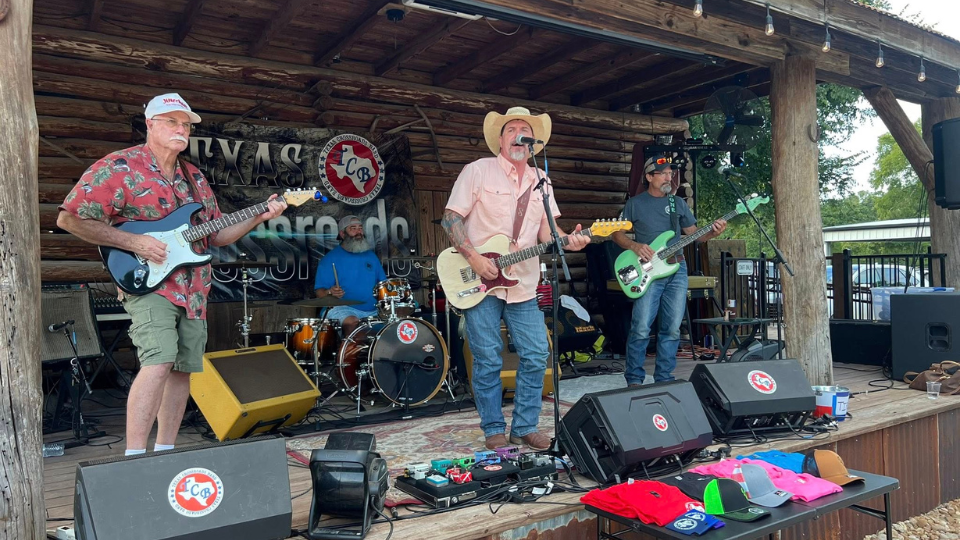 Live band performing outdoors on rustic stage, Texas theme, featuring guitars, drums, and colorful merchandise in foreground.