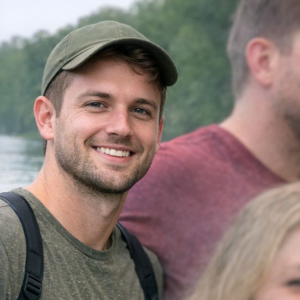 smiling man in outdoor setting wearing a green cap and casual attire nature background