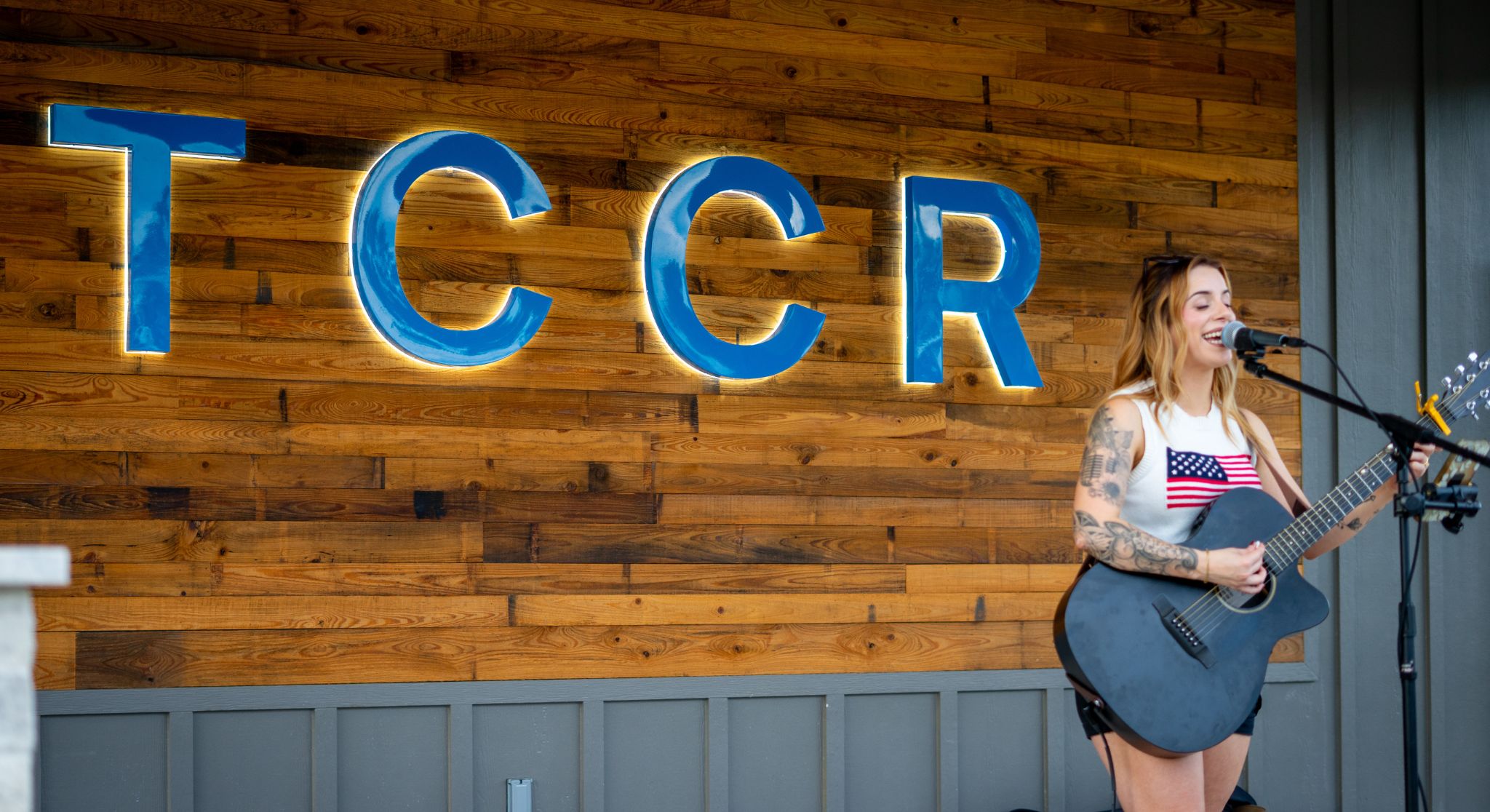 Generic music artist Female guitarist performs on a stage in front of a blue, backlit sign mounted on a wood-paneled wall.